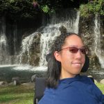 Photo of Sarah Cheung, who is sitting in her wheelchair in front of a waterfall. She is wearing sunglasses and has medium-length dark hair.
