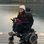 Photo of Julia Lamb. She is sitting in her power wheelchair on a dock in front of a lake and mountains. She is wearing a white toque and has long brown hair.