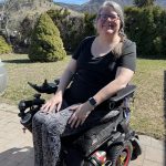 Photo of Michelle Hewitt. She is sitting in her power wheelchair in front of mountains and other picturesque nature. She has medium-length brown/gray hair and is wearing a black shirt and multicoloured leggings