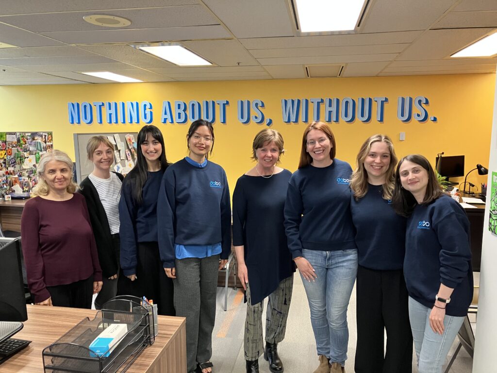 Minister Sheila Malcolmson with DABC staff in front of a blue and yellow mural that says "Nothing About Us, Without Us" at the DABC office. 